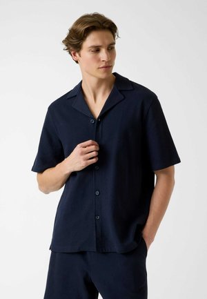 Junger Mann trägt ein marineblaues, kurzärmliges Hemd mit Kragen, schaut zur Seite, eine Hand an den Knöpfen, die andere in der Tasche.