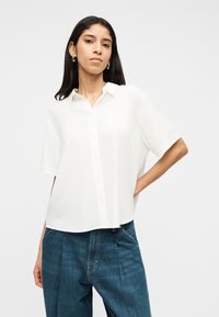 Γυναίκα με μακριά σκούρα μαλλιά, φορώντας ασημένια σκουλαρίκια, μια άνετη λευκή κοντομάνικη πουκάμισο με κουμπιά και μπλε τζιν ψηλόμεσα, στέκεται με το χέρι στη μέση.