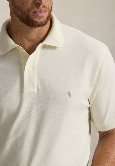 Šviesiai geltona polo marškinėliai iš tekstūruotos medžiagos, su trijų mygtukų užsegimu ir mažyčiu siuvinėtu logotipu kairėje krūtinės pusėje.