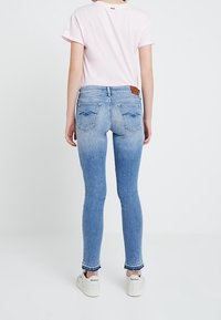 Camiseta rosa claro combinada con jeans skinny azules, mostrando un degradado de oscuro a claro. Presenta bolsillos traseros con detalle de costura y zapatillas blancas.