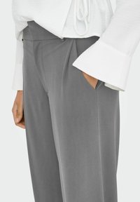 Sive odijeljene hlače glatke teksture, s visokim strukom, prednjim nabranim detaljima i bočnim džepovima, u kombinaciji s bijelom bluzom dugih rukava.