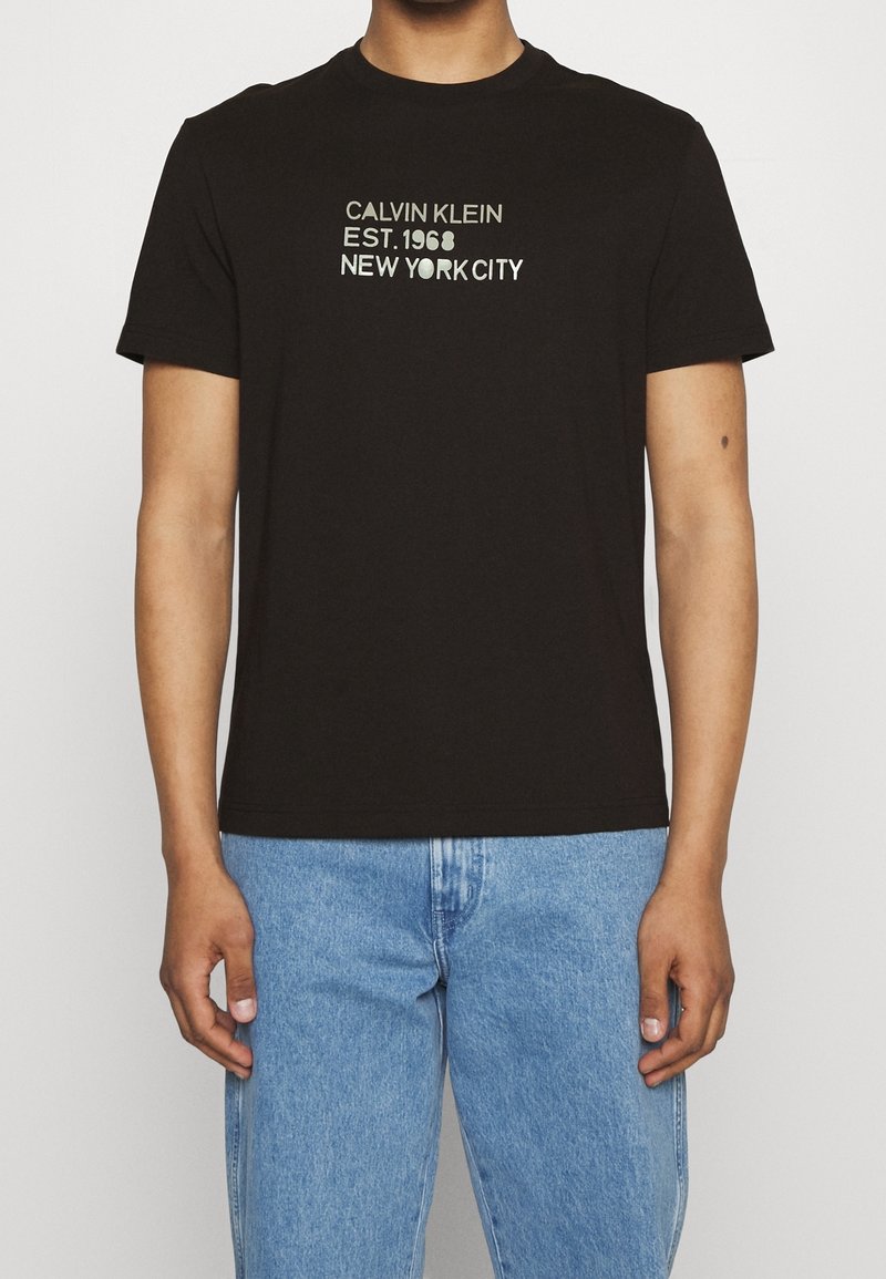 T-shirt en coton noir à manches courtes, avec un texte blanc : "CALVIN KLEIN EST. 1968 NEW YORK CITY" sur le devant. Conception ajustée.