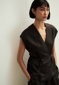Woman with short black hair wearing a sleeveless black wrap top with a tie belt, standing against a plain light wall.