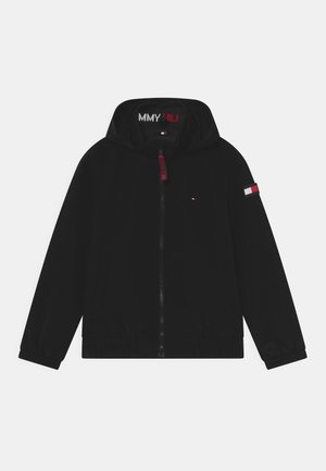 Veste noire à capuche zippée avec poignets élastiques, petit logo rouge, blanc et bleu sur la poitrine et la manche, et tirette de zip rouge.