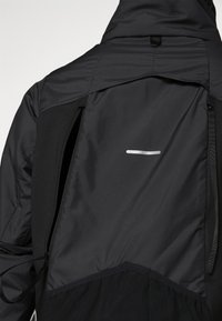 Dos de veste à capuche noire avec deux zips verticaux et une bande réfléchissante horizontale au centre du haut du dos.