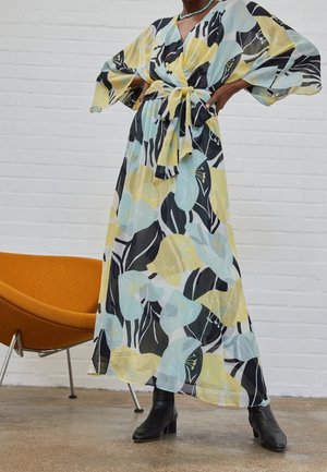 Femme portant une longue robe portefeuille à fleurs avec des manches larges et une taille nouée, associée à des bottines noires à talons, debout près d'une chaise orange.