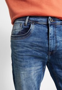 Blå denim jeans med en falmet tekstur, med en frontlomme og en metalknaplukking. Accepteres af en orange trøje ved taljen.