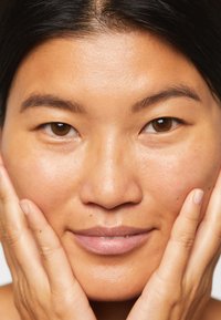 Gros plan sur un visage avec une peau lisse et brun clair. L'individu a des yeux brun foncé et ses mains entourent doucement ses joues. Un éclairage naturel met en valeur la texture de la peau.