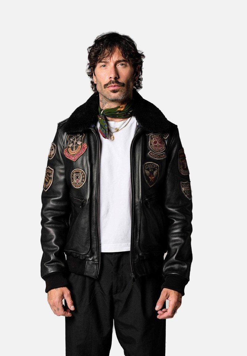 Veste bomber en cuir noir avec un col en shearling, ornée de plusieurs patchs brodés sur les manches. Portée par-dessus un t-shirt blanc.