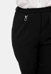 Schwarze Hose mit elastischem Bund, ausgestattet mit einem silbernen Metallakzent und Seitentaschen, aus einem glatten, strukturierten Stoff gefertigt.