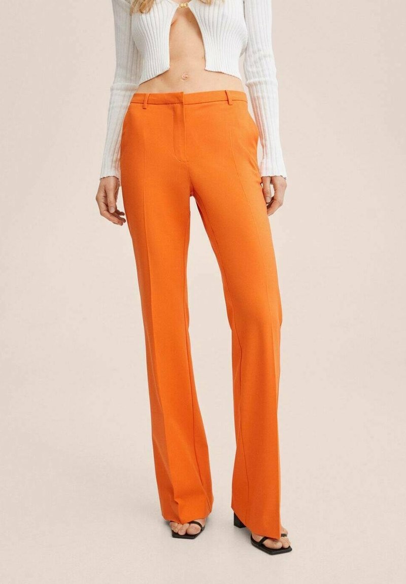 Femme portant un pantalon droit orange vif associé à un top court blanc côtelé et des talons ouverts noirs sur un fond beige.
