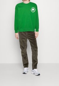 Sudadera verde con un escote redondo y detalle de logotipo blanco en la manga, combinada con pantalones de pana oliva y zapatillas deportivas.