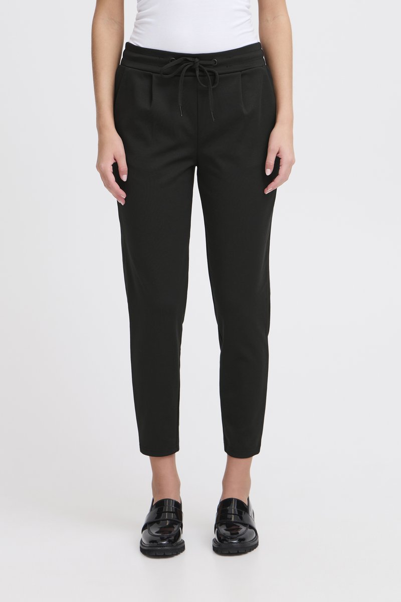 Fekete, enyhén szűkülő nadrág rugalmas derékpánttal és húzózsinórral, elöl behúzásos részletekkel és sima tapintású anyagból. Fényes fekete cipővel viselve.