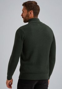 Dunkelgrüner, strukturierten Pullover mit hohen Kragen und gerippten Bündchen. Mit einem Rautenmuster auf dem Rücken und einem kleinen Logo in der Nähe des Kragens.