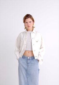 Bela denimska jakna z velikimi gumbi, kombinirana s svetlo rebrasto majico in visokim denimsko krilom v zbledeli modri barvi. Gladek, sproščen videz.