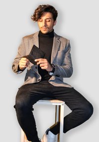 Man with curly hair seated, wearing patterned blazer and black turtleneck, holding and looking at an open black wallet.