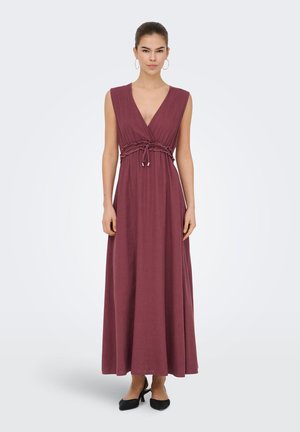 Žena stojaca, oblečená v bordových bez rukávov dlhých šatách s hlbokým výstrihom do V, s nariaseným pásom previazaným vpredu a čiernymi špicatými podpätkami.