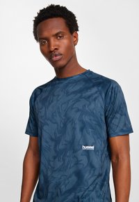 Man med kort lockigt hår som bär en blå mönstrad Hummel Performance kortärmad träningströja mot en enfärgad ljus bakgrund.