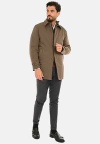 Giacca impermeabile marrone con colletto, chiusura con bottoni e tasche laterali; abbinata a pantaloni grigi e mocassini neri.