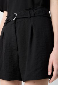 Black shorts made from textured material, featuring a wide belt with a silver buckle, front pleats, and side pockets.