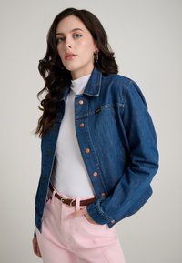 Tamsiai mėlyna džinsinė striukė su vario spalvos spaudėmis, klasikinio apykaklės ir šoninių kišenių, dėvima ant balto viršaus ir rožinių kelnių.