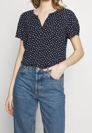 Vrouw die een marineblauwe korte-mouwen blouse draagt met witte en lichtblauwe patronen en een hoge taille blauwe spijkerbroek met haar hand in de zak.
