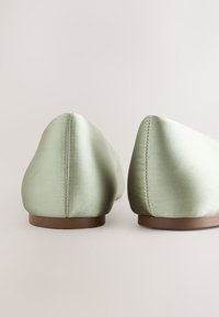 Paire de ballerines en satin vert clair vues de dos, avec semelles plates marron sur fond neutre.