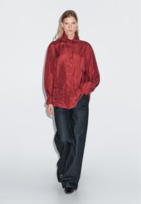 Red satin shirt with a high collar and button closure, paired with dark denim wide-leg pants. Smooth texture, relaxed fit, and minimal design.