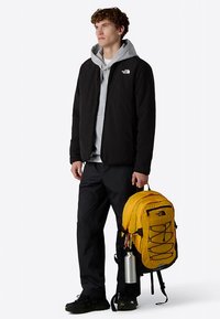 Must jakk halli kapuutsiga, mustad püksid ja kollane seljakott. Seljakotil on kinnitusköied ja veepudeli hoidik.