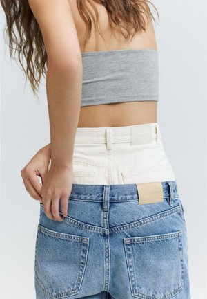 Woman wearing layered high-waist pants: white denim underneath and loose blue denim on top, paired with a grey tube top.