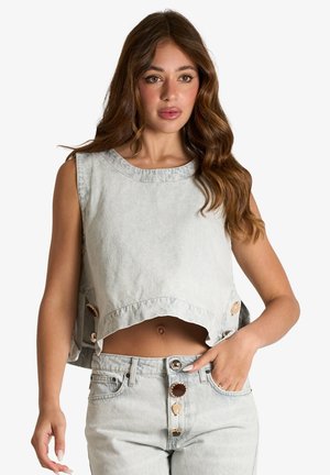 Donna con capelli lunghi e mossi che indossa un top corto senza maniche in denim chiaro e jeans a vita alta abbinati con bottoni decorativi.