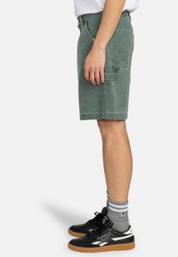 Shorts en denim verts avec une coupe droite et des poches latérales, associés à des baskets noires et des chaussettes grises côtelées.