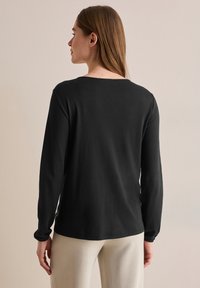 Schwarzes Langarmshirt mit rundem Ausschnitt und weicher Textur, kombiniert mit hellen Hosen. Ansicht von hinten, die ein einfaches Design zeigt.