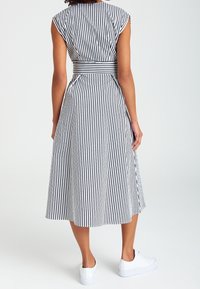 Robe rayée en noir et blanc, sans manches, taille cintrée avec une ceinture, jupe fluide jusqu'au milieu du mollet, portée avec des baskets blanches.