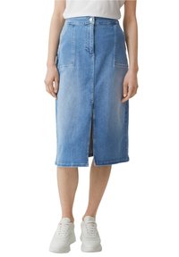 Kobieta ubrana w niebieską dżinsową spódnicę do kolan z przednim rozcięciem, kieszeniami naszytymi i białymi trampkami, skróconą w talii i nogawkach.