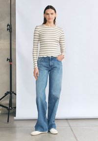 Pasiasty, długi rękaw top w kolorze kremowym i granatowym, zestawiony z jasnoniebieskimi, rozszerzanymi dżinsami. Modelka stoi na tle neutralnym. Widać casualowe obuwie.