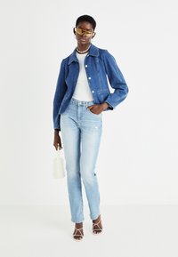 Chaqueta de mezclilla en azul oscuro con cuello, camiseta blanca con canalé, pantalones vaqueros azul claro y tacones blancos con tiras. Los accesorios incluyen unas gafas de sol amarillas y un bolso blanco.