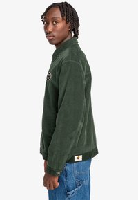 Grøn kordjakke med krave, prydet med et patch-logo, knapmanchetter og sidelommer. Bæres sammen med blå jeans.