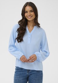 Frau mit langen, welligen Haaren, die eine hellblaue Langarmbluse mit Knöpfen und blaue Jeans trägt, lächelt mit sanft gefalteten Händen.