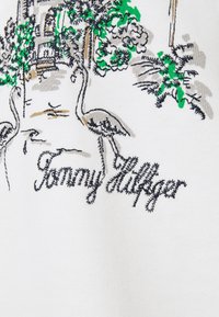 Tela blanca con bordado colorido de follaje, dos flamencos grises y el texto "Tommy Hilfiger" en hilo negro.