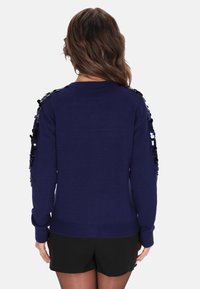 Donna dai capelli castani mossi che indossa un maglione blu navy con paillettes nere e argento sulle maniche e pantaloni corti neri, rivolta di spalle rispetto alla macchina fotografica.