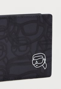 Mustra tekstuuriga must rahakott, millel on prillide kontuuride disain, paremas alumises nurgas on silmatorkav valge logo inimesest, kes kannab prille.