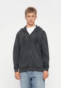 Young man wearing a dark grey zipped hoodie with "GAP" logo and light blue jeans, standing against a plain white background.