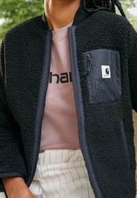 Tekonahka musta takki vetoketjullisilla taskuilla, jossa on sileä kangaspäällyste. Sen alla vaalea vaaleanpunainen paita mustalla logotekstillä.