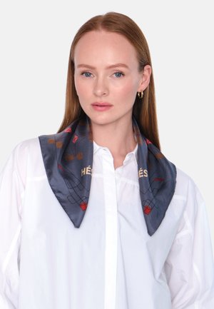 Silkkihuivi tummansinisellä pohjalla, jossa on geometrinen kuvio punaisilla ja kultaisilla yksityiskohdilla. Kolmiomainen muoto, sileä rakenne, asetettuna valkoisen paidan päälle.