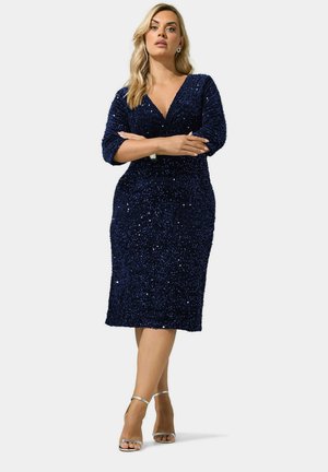 Femme aux formes généreuses debout, bras croisés, portant une robe midi bleu marine à sequins et des sandales à talons hauts argentées sur fond blanc.