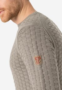 Grauer Strickpullover mit strukturiertem Muster, Rippdetails und einem kleinen Logopatch auf der linken Schulter.