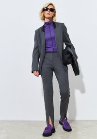 Harmaat räätälöity puku tummalla kauluksella, violetti korkea paita ja violetit loafersit. Housuissa on etuleikkaukset; asusteena mustat aurinkolasit.