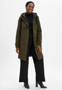 Smiling woman wearing a long olive green hooded coat, black turtleneck, black wide-leg pants, and black heeled boots standing on white background.