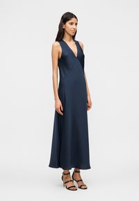 Γυναίκα με αμάνικο μάξι φόρεμα από ναυτικό μπλε σατέν με V λαιμόκοψη, σετ με μαύρα ψηλοτάκουνα σανδάλια με λουράκια, στέκεται μπροστά σε απλό φόντο.
